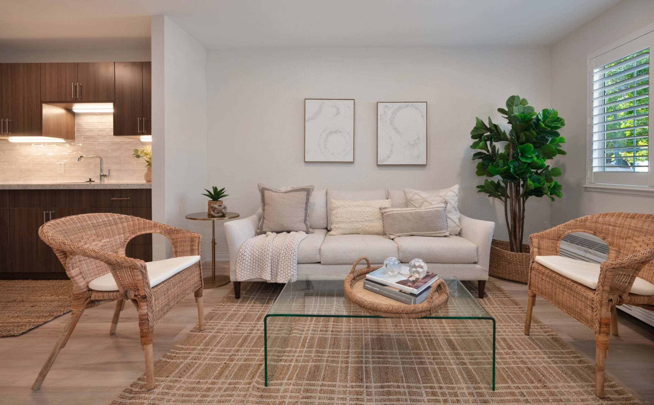 A modern assisted living living room with a white sofa, two wicker chairs, a glass coffee table, neutral decor, plants, and a view of a stylish kitchen in the background. Large window with shutters lets in natural light.